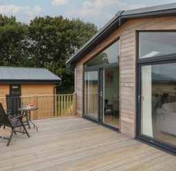 A wooden deck with two black chairs and a small round table outside a wooden house with large glass doors at Cove Lodge in Ilfracombe