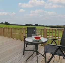 A wooden deck with a round glass table and two black chairs overlooking green fields at Cove Lodge in Ilfracombe