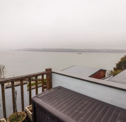 A balcony with a wooden railing overlooking a body of water with hills and buildings in the distance at Cheriton in Pembroke Dock