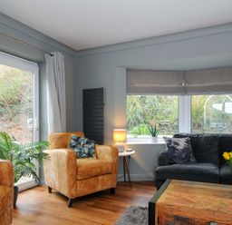 A living room with armchairs a sofa coffee table and windows overlooking plants at Waves Edge in Looe