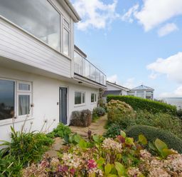 An exterior view of a white house with large windows a garden path and shrubs by the sea at Waves Edge in Looe