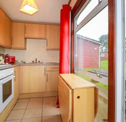 A kitchen with wooden cabinets a window with red curtains and a foldable wooden table near a picnic bench outside at Pepper Cove near St Merryn