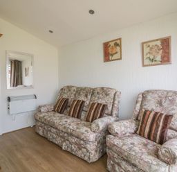 A living room with patterned sofa and armchair with striped cushions paintings on the wall and a wall-mounted heater at Warren Cove near St Merryn