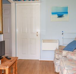 A room with a floral patterned sofa with blue cushions a wooden side table with a TV and remote control a white door a wall heater and a painting of a boat on a beach at High Cove near St Merryn