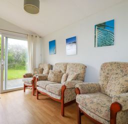 A sitting room with floral armchairs and sofa and sliding glass door opening to a garden at Porthcothan Bay in St Merryn
