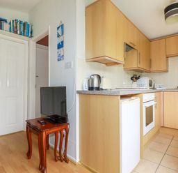 A kitchen with wooden cabinets a countertop with a kettle toaster and microwave a small fridge an oven a small wooden table with a television and books on a shelf above a door at Porthcothan Bay in St Merryn