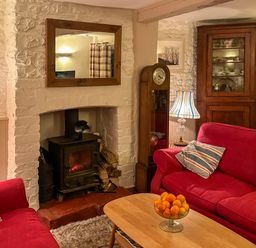 A living room with red sofas a wooden coffee table with a bowl of oranges a fireplace with a wood stove and a tall clock at Lamorna Cottage in Chipping Norton