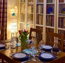 A dining table set with plates glasses and cutlery beside a bookshelf with glass doors and a lamp with floral design at Lamorna Cottage in Chipping Norton