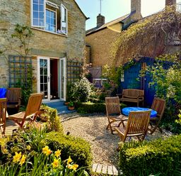 An outdoor garden area with wooden chairs around a blue round table and a wooden bench near a stone house and a blue shed