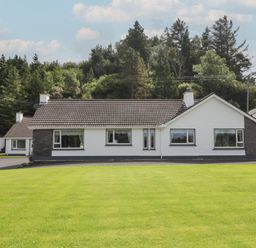 A single-story white house with a tiled roof and multiple windows in front of a lawn and trees at Nelly's House in Gortahork County Donegal