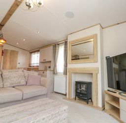 A living room with a beige sofa a patterned ottoman a small electric stove in a wooden frame a wall mirror curtains and a flat screen TV on a stand at Drake 11 in Cayton Bay