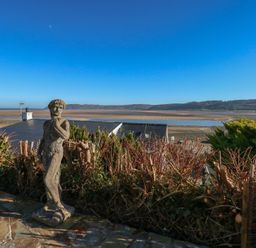 A garden patio with a stone statue overlooking rooftops hills and a body of water at Swn Y Wylan in Red Wharf Bay near Benllech