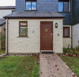 A front entrance with a brown door and window on a cream textured wall with a path and grass in front at Coombe Cottage in Falmouth