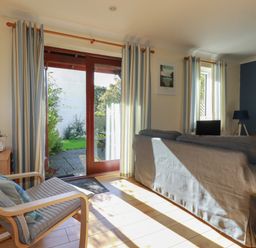 A living room with a gray sofa, a wooden armchair with cushions, striped curtains, a floor lamp, and glass doors opening to a garden at Coombe Cottage in Falmouth