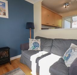 A living room with a grey sofa and two decorative pillows by a blue wall with a framed picture and a small black stove at Coombe Cottage in Falmouth