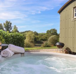 An outdoor hot tub with rolled towels on the edge a small round table with drinks and flowers a metal chair a green corrugated metal building a charcoal grill and trees in the background at Shepherds Joy Kennerleigh near Cheriton Fitzpaine
