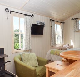 A small room with a wood stove a green armchair a bed with towels on it and a wall-mounted TV at Shepherds Joy in Kennerleigh near Cheriton Fitzpaine