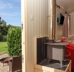 A small room with a wood stove a mounted TV a bed with patterned bedding and windows with curtains at Shepherds Bliss in Kennerleigh near Cheriton Fitzpaine