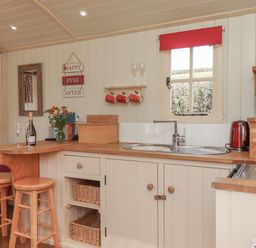 A kitchen with a wooden countertop a sink two bar stools a window with a red blind and kitchenware at Shepherds Bliss in Kennerleigh near Cheriton Fitzpaine