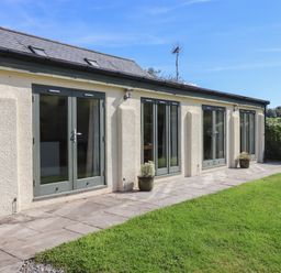 A cream colored building with multiple green framed windows and doors and two potted plants on a stone patio with grass surrounding at Binneford Barn in Kennerleigh near Cheriton Fitzpaine