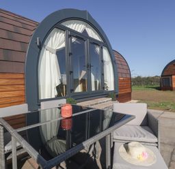 An outdoor patio with a glass table and cushioned chairs in front of a curved wooden cabin at Leadon View Oak in Upleadon near Newent