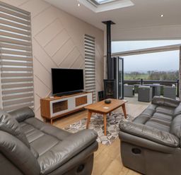 A living area with two grey leather sofas a wooden coffee table a flat screen tv on a wooden stand and an open sliding door leading to an outdoor seating area with views of trees and fields at The Lodge @ Racecourse House in Babell near Holywell