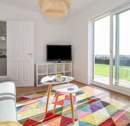 A living room with a gray sofa colorful geometric rug small white tables flat screen tv on a white shelf and sliding glass door to a garden at Beachcomber Maer near Bude