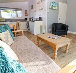 An open living room and kitchen area with a beige sofa with blue and beige cushions a wooden coffee table a black armchair and a small dining table with wooden chairs at Chalet 3 Wipeout in Hayle