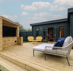 An outdoor wooden deck with a television, two yellow chairs and table with drinks and a lounge chair with cushions at Badger's Burrow in South Luffenham near Edith Weston