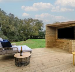 An outdoor wooden deck with a sofa, round table with a bottle and glasses, and a built-in wooden TV unit at Badger's Burrow in South Luffenham near Edith Weston