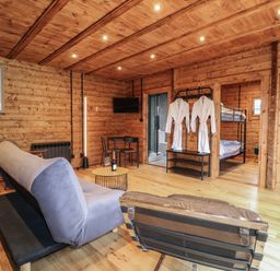 A living area with wooden walls and ceiling containing a couch, chair, round table with wine, TV on wall, two white robes hanging and visible bedrooms at Badger's Burrow South Luffenham near Edith Weston