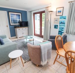 A living room with a blue sofa gray rug glass coffee table and wooden dining table with chairs at Chy Lowena in Goldenbank near Budock Water