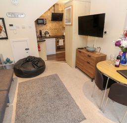 A living room with sofa bean bag table chairs flowers and TV and view into kitchen at 5 Lyonesse Apartments in St Ives