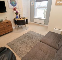 A living room with a gray sofa a wooden cabinet a small dining table with chairs and a wall-mounted television at 5 Lyonesse Apartments in St Ives