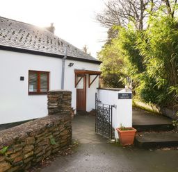 A small white cottage with a brown door and window behind a stone wall and metal gate surrounded by greenery at Artists Cottage in Minffordd near Penrhyndeudraeth