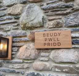 A stone wall with a wooden sign reading Beudy Pwll Pridd and a lit wall-mounted lantern at Beudy Pwll Pridd in Llangollen
