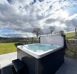 A hot tub on a paved patio next to a stone wall with grass and hills in the background at Beudy Pwll Pridd in Llangollen