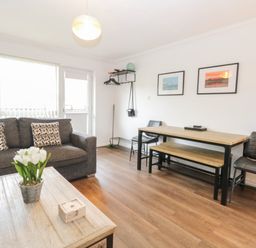 A living area with a dark sofa with patterned cushions a wooden coffee table with flowers a wooden dining table a bench two chairs and wall art at 102 Waterside Park in Corton