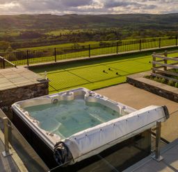 An outdoor hot tub on a patio next to a fenced green lawn with bocce balls and a view of rolling hills at High View Barn in Sedbergh