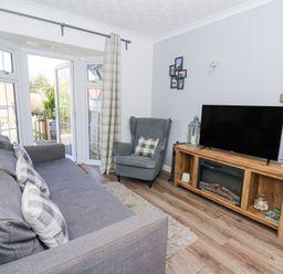 A living room with a gray sofa and armchair a wooden media unit with a TV and a bay window with doors and curtains at The Cwtch in Aberporth