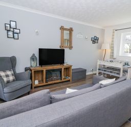 A living room with a gray armchair a sofa a wooden TV stand with a TV and a fireplace a dining table and window at The Cwtch in Aberporth