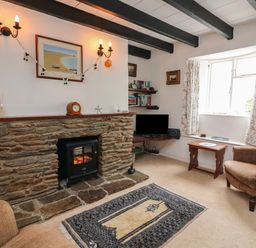 A living room with a stone fireplace a small stove an armchair a wooden table a patterned rug and a window with floral curtains at Vine Cottage in East Prawle
