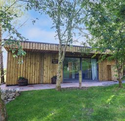 A wooden lodge with large glass doors surrounded by trees and grass at Orchard Lodge in Maerdy near Corwen