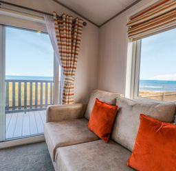 A living area with a beige sofa and orange cushions next to sliding glass doors and windows overlooking a balcony and the sea at Seaview Lodge in Bacton