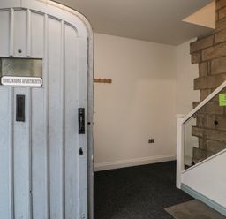 An entrance hall with a closed door labeled Tomlinsons Apartments a coat rack on the wall and stairs with glass panels and stone wall at The Coquet Nest in Rothbury
