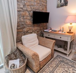 A wicker armchair with cushions next to a basket with a blanket and a dog bowl beside a stone wall a console table with books and a lamp on top and a wall-mounted TV above it at The Coquet Nest in Rothbury