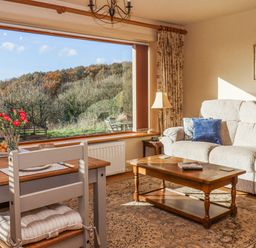 A living room with a sofa, coffee table, dining table with chairs, and a large window showing an outdoor view at Sherway Dairy in Clyst Hydon near Bradninch