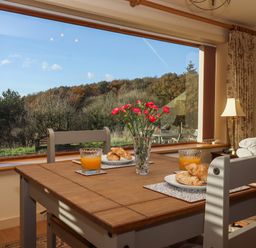 A dining area with a wooden table set with croissants and orange juice next to a large window showing trees outside at Sherway Dairy in Clyst Hydon near Bradninch