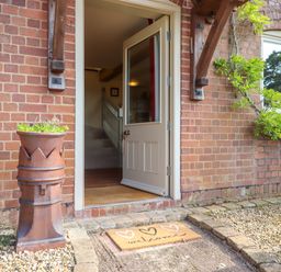 An open front door with a welcome mat and a plant pot outside a brick house at Sherway Shippen in Clyst Hydon near Bradninch
