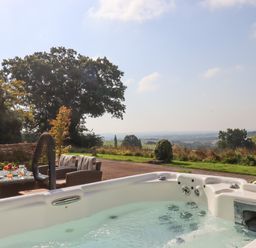 An outdoor hot tub with water jets near a table with chairs and drinks overlooking a countryside landscape at Sherway Shippen in Clyst Hydon near Bradninch